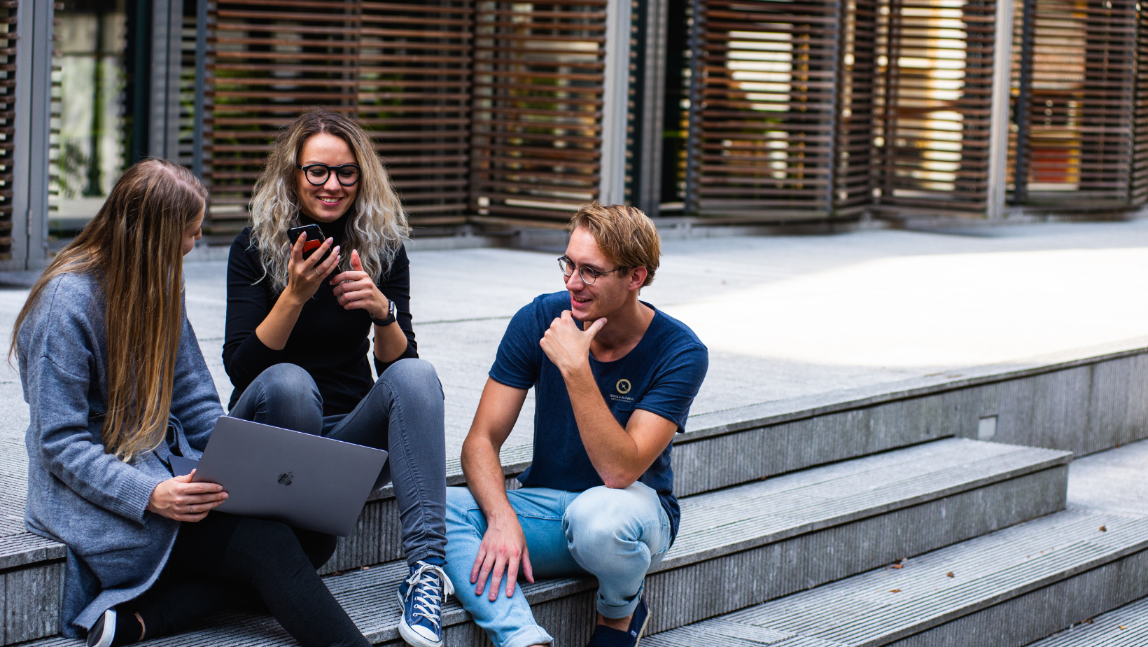 Obrazek dekoracyjny pełniący funkcję przycisku przekierowującego do zakładki z informacjami o studiach licencjackich. Na zdjęciu trzech studentów siedzi na schodach i rozmawia. Studentka trzyma w ręku laptopa.