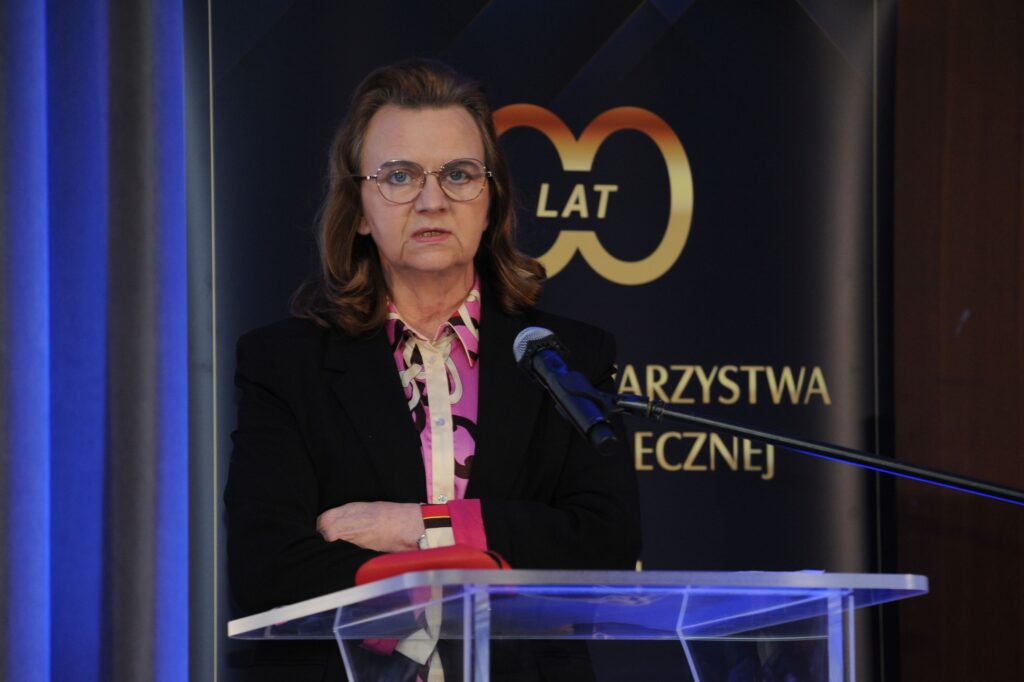 Photo presents: on the stage behind the lectern, Prof. Gertrude Uścińska stands and speaks in front of the microphone. The logo of the event is visible in the background.