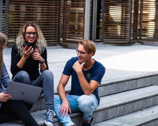 Obrazek dekoracyjny pełniący funkcję przycisku przekierowującego do zakładki z informacjami o studiach licencjackich. Na zdjęciu trzech studentów siedzi na schodach i rozmawia. Studentka trzyma w ręku laptopa.